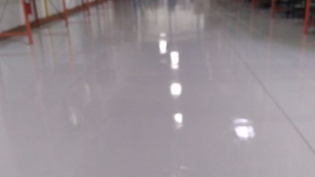 Polished epoxy warehouse floor reflecting overhead lights and red support posts along the edges, creating a glossy, uniform surface.
